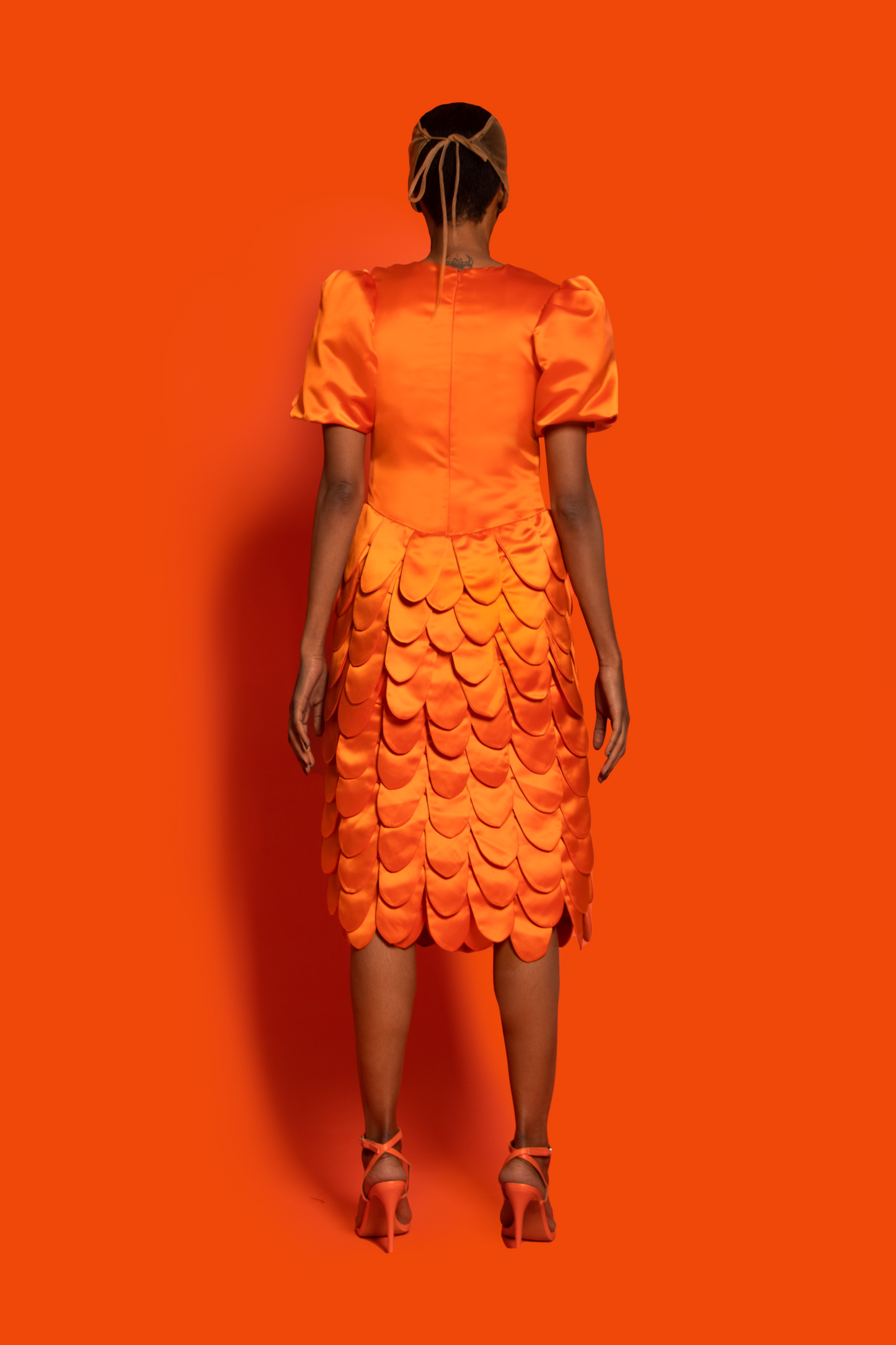 Back view. An orange silk taffeta midi dress features a puffy sleeve, dropped waist, and round shapes layers finished with silk lining.