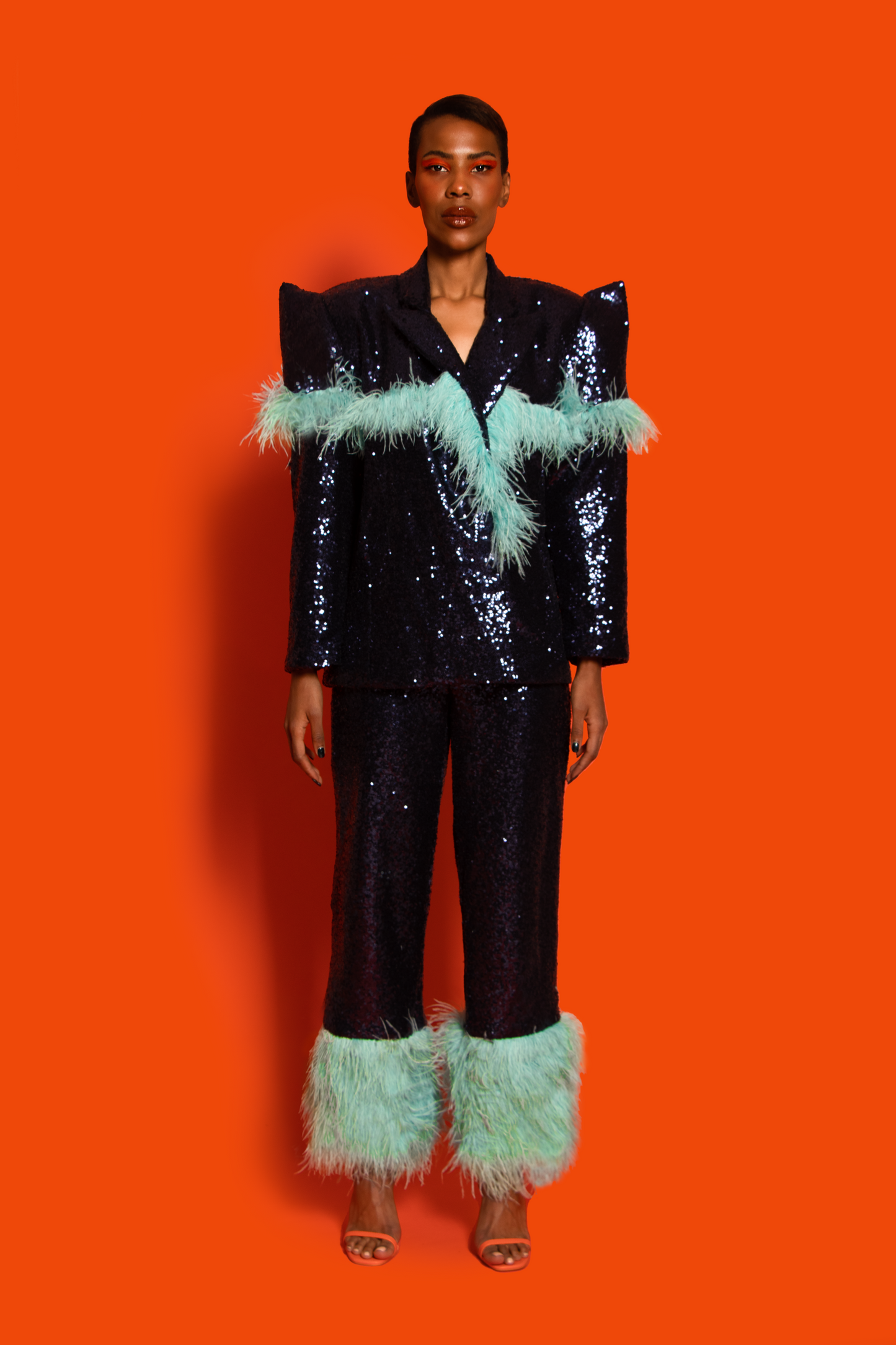 Front View. A navy sequin embellished constructed bold shoulder suit blazer features pop blue feathers on the chest and sleeve lined with navy taffeta.